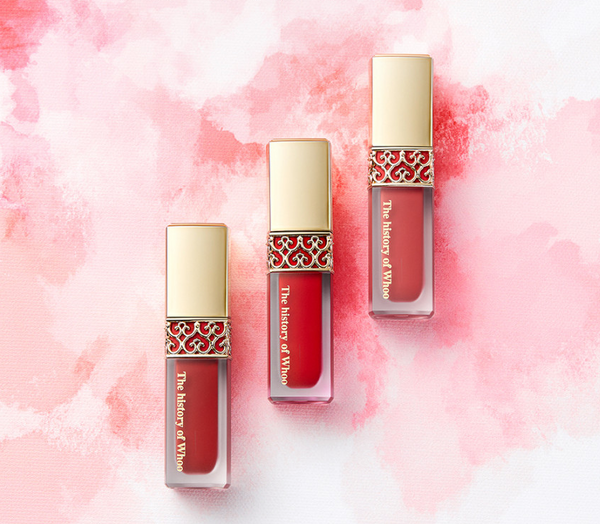 Three lip glosses with decorative caps on a pink and white watercolor background